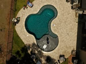 High Angle Drone View Of A Kidney-Shaped Backyard Swimming Pool With Dark Plaster, A Wide Tanning Ledge, And Tan Paver Decking Surrounded By A Black Fence And Ocala Pool Remodeling.