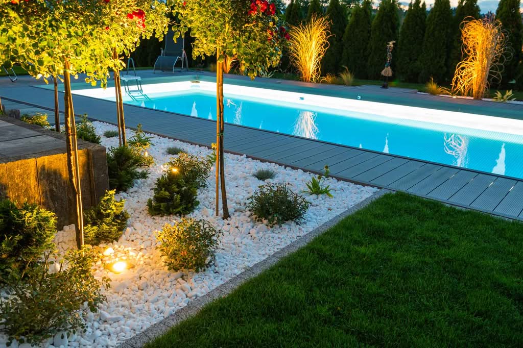 Equipment Manuals 3 Modern Pool, Narrow Lap Swimming Pool At Night With Bright Blue Underwater Led Lighting, Surrounded By A Gray Composite Deck, Illuminated Landscaping With White River Rock, And Manicured Trees.