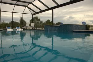 Water-Level View Of A Swimming Pool Inside A Black Screen Enclosure, Featuring A Raised Blue Mosaic Tile Water Wall With Small Waterfalls And Submerged White Lounge Chairs Against A Cloudy Sky.