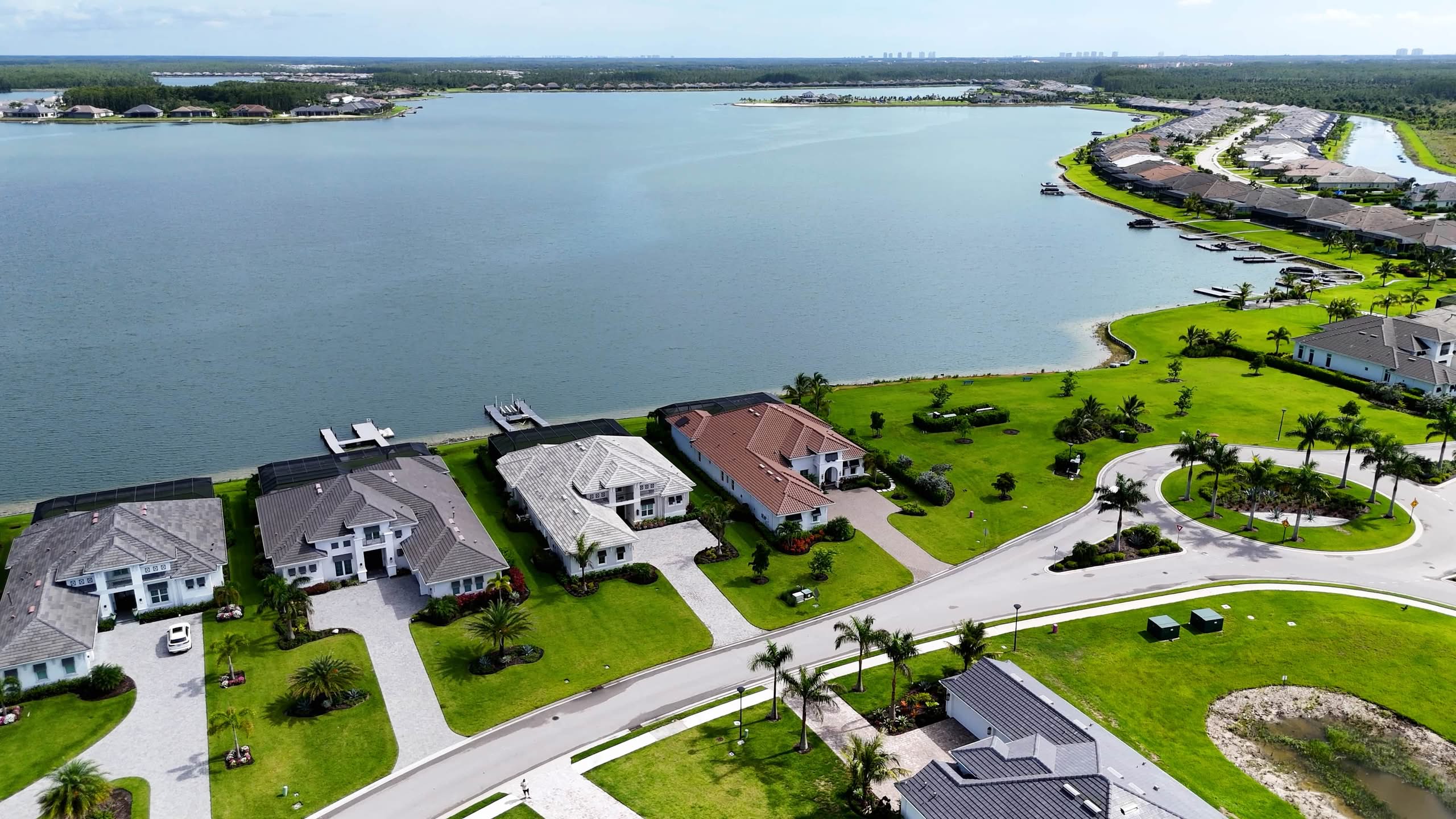 Saltwater Conversions in The Villages 1 High-Angle Drone Photograph Of A Row Of Modern Luxury White-Walled Mansions With Grey Roofs Bordering A Large Calm Lake With Private Docks And Manicured Green Lawns.