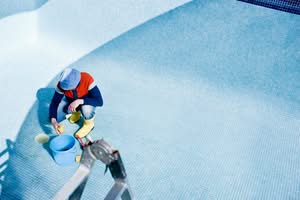 A Worker Wearing Yellow Boots And A Hat Crouches In An Empty, Blue-Tiled Swimming Pool, Using A Sponge And A Blue Bucket To Clean The Tiles Manually With Veteran-Led Precision.