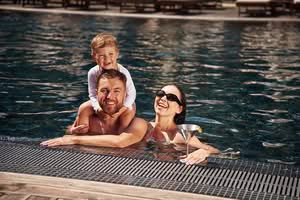 A Smiling Father With A Young Son On His Shoulders And A Mother Holding A Cocktail, Enjoying A Sunny Day In A Luxury Outdoor Swimming Pool For The Best Pool Heater For The Villages In Florida.