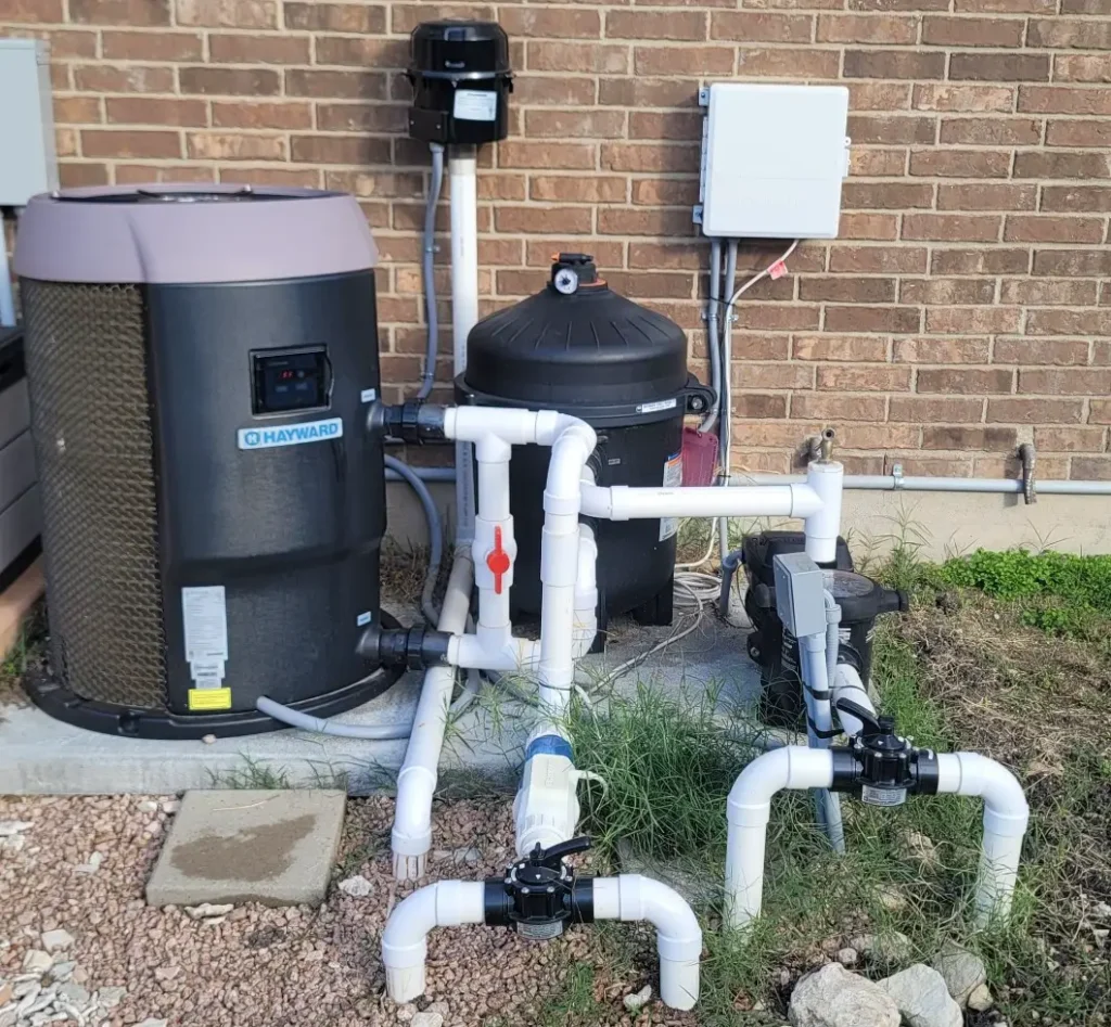 Best Pool Heater for The Villages in Florida Climate 3 A Professional Residential Pool Equipment Pad Installation Featuring A Hayward Heatpro Heat Pump, A Black Cartridge Filter, And Pvc Plumbing With Jandy Valves Against A Brick Exterior Wall.