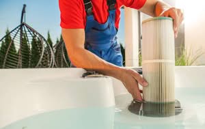 Close-Up Of A Professional Filter Cleaning In Blue Overalls Inserting A New White Pleated Cartridge Filter Into The Filter Housing Of A Hot Tub Filled With Clear Water.