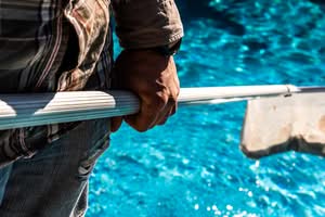 A Pool Maintenance Technician Using A Leaf Skimmer Rake To Clear Debris From A Residential Swimming Pool And Last-Minute Pool Inspections In Orlando, Florida.