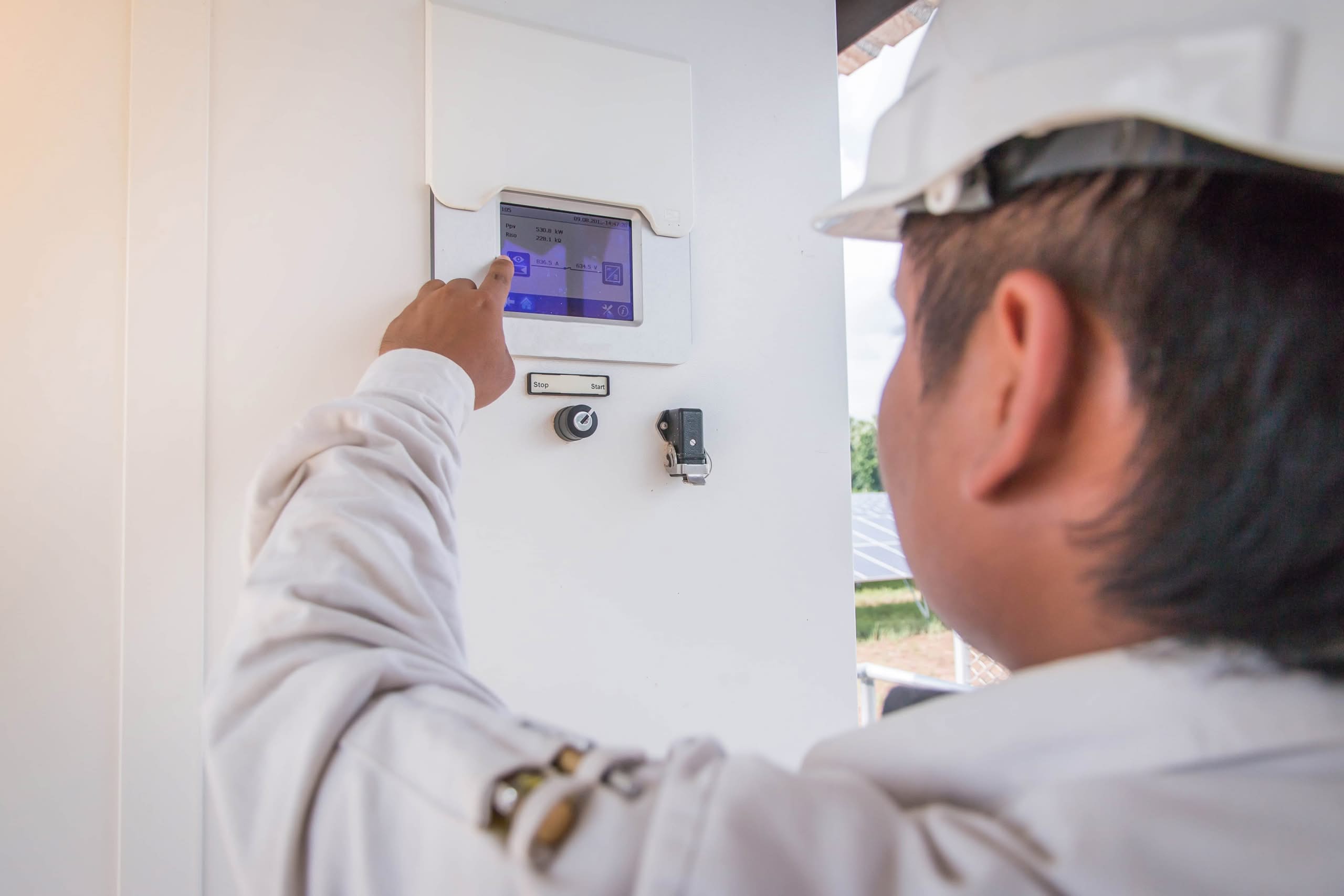 Close-Up View Of A Clean Energy Technician Wearing A White Hard Hat And Work Jacket, Pointing At And Interacting With A Touchscreen Human Machine Interface (Hmi) Panel For A Solar Power Inverter System At A Solar Farm Facility.