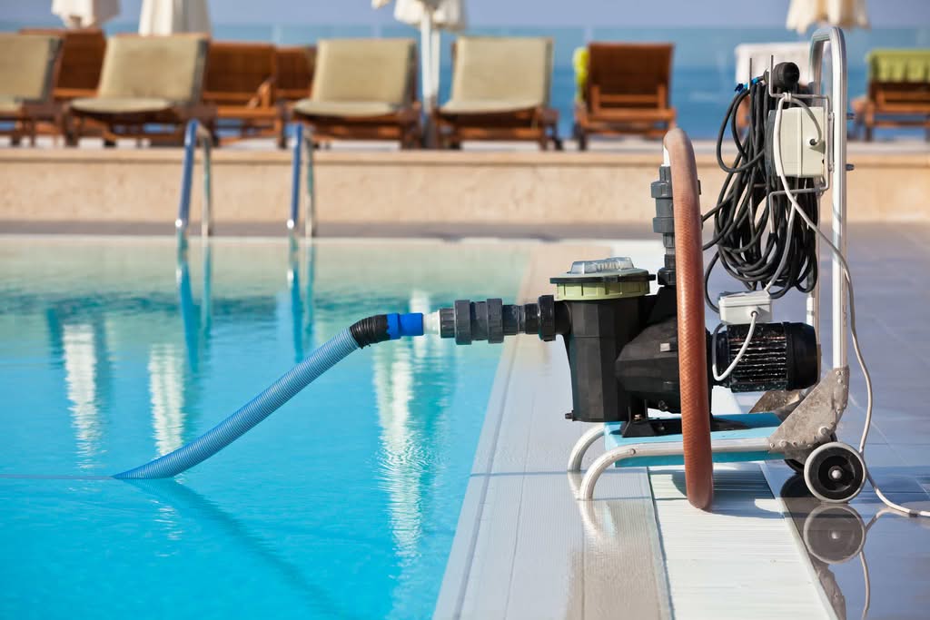 A Professional Portable Commercial Pool Maintenance Pump And Vacuum Cart System Operating On The Edge Of A Large Resort Swimming Pool With Lounge Chairs In Miami, Florida.