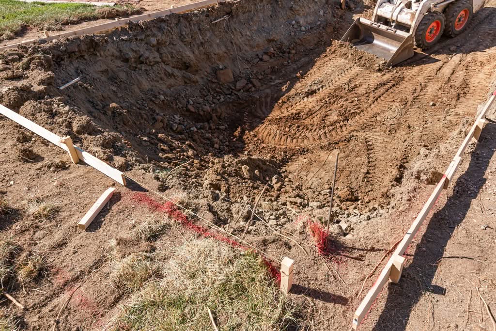 A Split View Showing The Contrast Of A Residential Pool Project: A Small Skid Steer Bulldozer Excavating Dirt In A Marked-Off Backyard Vs. A Finished High-End Geometric Swimming Pool With Crystal Blue Water, Stone Pavers, And Modern Lounge Chairs Under A Blue Umbrella In Phoenix, Arizona.