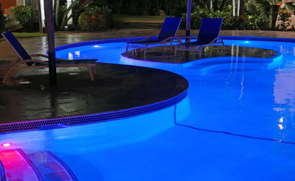 A Luxury Backyard Swimming Pool Beautifully Illuminated At Night With Vibrant Blue Led Pool Lights Installation And Lounge Chairs On A Paved Deck In Miami, Florida.