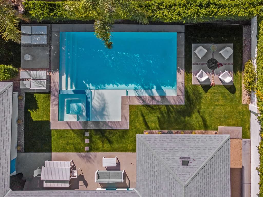 A Top-Down Aerial View Of A Luxury Pool Renovation Featuring Curved Light Blue Mosaic Tiles, Custom Stone Coping, A Grey Paver Deck, And Modern Striped Lounge Chairs Shaded By A Vibrant Blue Umbrella In Palm Springs, California.