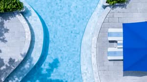 A Top-Down View Of A Luxury Minimalist Swimming Pool Featuring Clean Geometric Lines, Light Blue Mosaic Tiles, And A Natural Stone Paver Deck With Modern Loungers And A Vibrant Blue Umbrella In Scottsdale, Arizona.