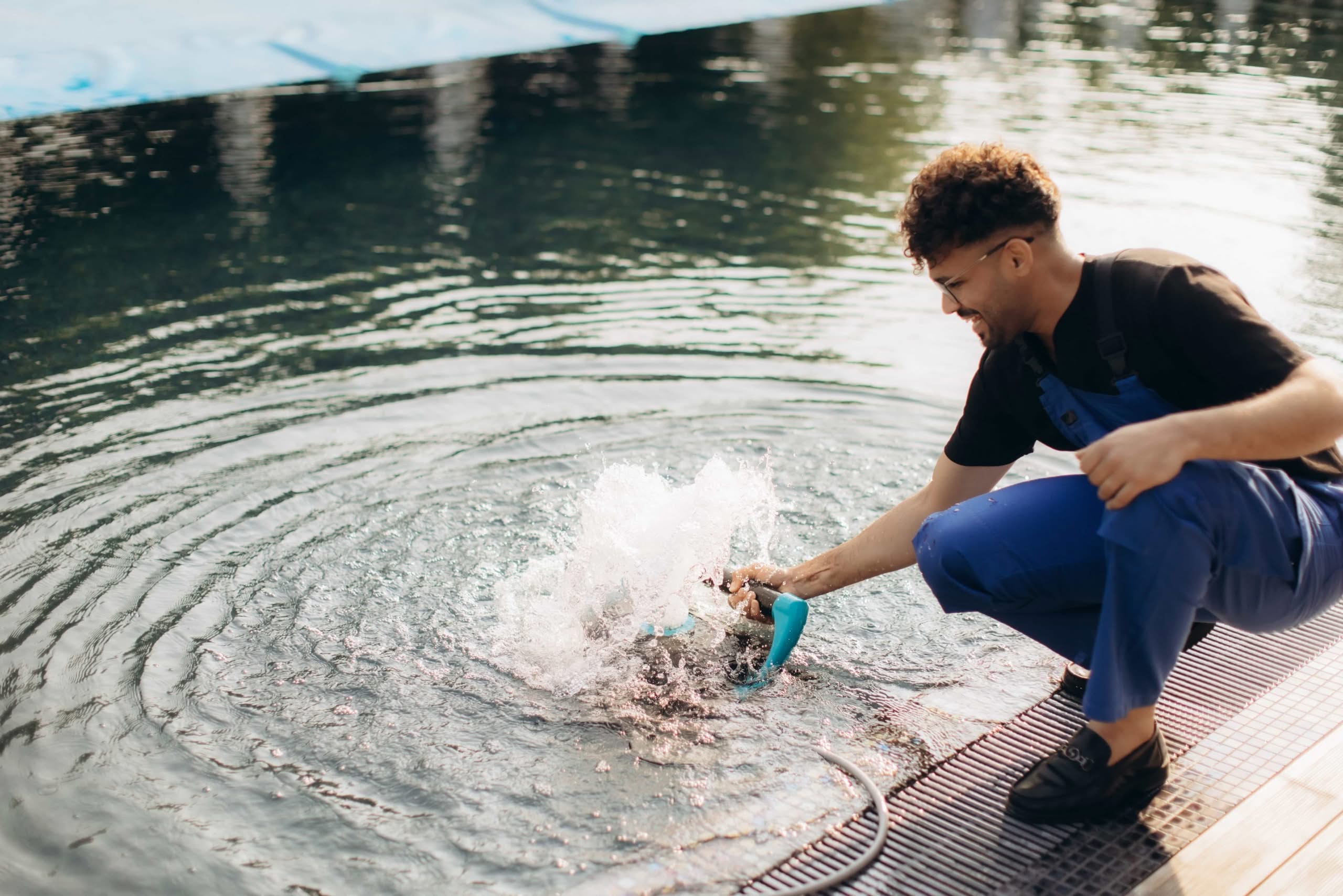A Pool Maintenance Worker Wearing Blue Overalls Crouches At The Edge Of A Swimming Pool Using A Cordless Cleaning Tool That Creates A Splash In The Water, Illustrating Troubleshooting For High Pool Filter Pressure In Las Vegas, Nv.