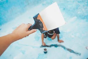 A Hand Holds Up A Wide Putty Knife Scraper In The Foreground While A Pool Technician Works On Repairing A Long Crack In The Bottom Of An Empty Blue Swimming Pool, Showcasing Professional Pool Resurfacing Options In Miami, Fl.