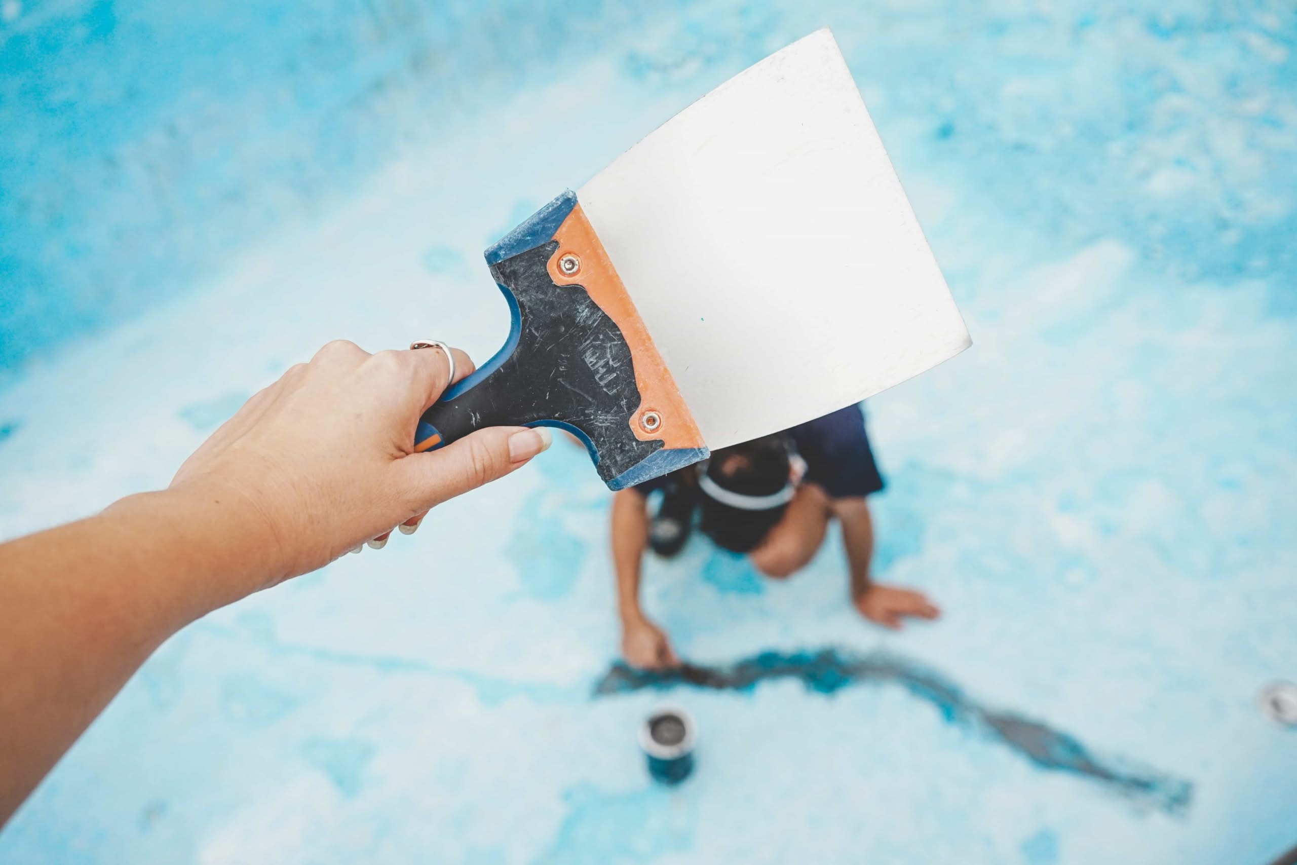 A Hand Holds Up A Wide Putty Knife Scraper In The Foreground While A Pool Technician Works On Repairing A Long Crack In The Bottom Of An Empty Blue Swimming Pool, Showcasing Professional Pool Resurfacing Options In Miami, Fl.